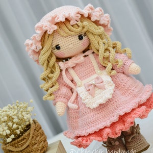 Pode incluir: Uma boneca de croch&ecirc; com cabelo loiro encaracolado, vestindo um vestido rosa com um avental branco e um la&ccedil;o rosa. A boneca est&aacute; usando sapatos marrons e um chap&eacute;u rosa de croch&ecirc;.