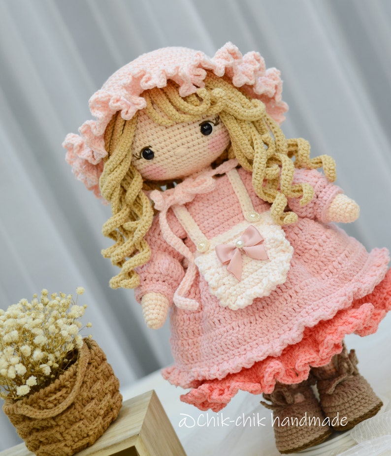 Pode incluir: Uma boneca de croch&ecirc; com cabelo loiro encaracolado, vestindo um vestido rosa com um avental branco e um la&ccedil;o rosa. A boneca est&aacute; usando sapatos marrons e um chap&eacute;u rosa de croch&ecirc;.