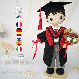 May include: A crochet doll dressed in a black graduation gown with a red stole, a black mortarboard cap with a red tassel, and grey trousers. The doll holds a white diploma tied with a red ribbon and a small bouquet of red and white flowers.