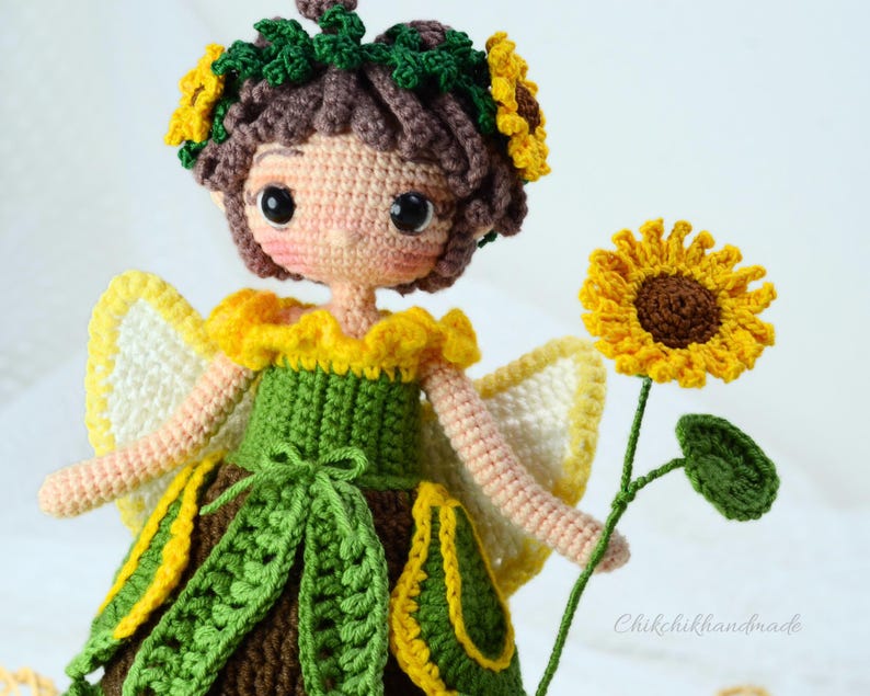 May include: A crocheted sunflower fairy doll with a green dress, yellow and white wings, and a sunflower in her hand. The doll has brown hair and a sunflower crown.