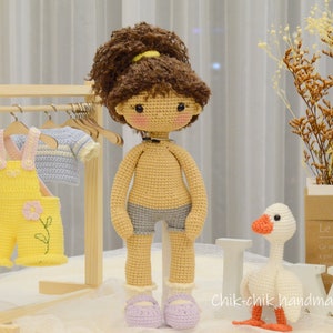 May include: A crocheted doll with brown hair and a blue and white outfit stands next to a crocheted goose. The doll is wearing a necklace and is holding a small wooden hanger. The goose is standing on a wooden block with the words "Chik-chik handmade" written on it.