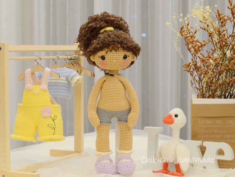 May include: A crocheted doll with brown hair and a blue and white outfit stands next to a crocheted goose. The doll is wearing a necklace and is holding a small wooden hanger. The goose is standing on a wooden block with the words "Chik-chik handmade" written on it.