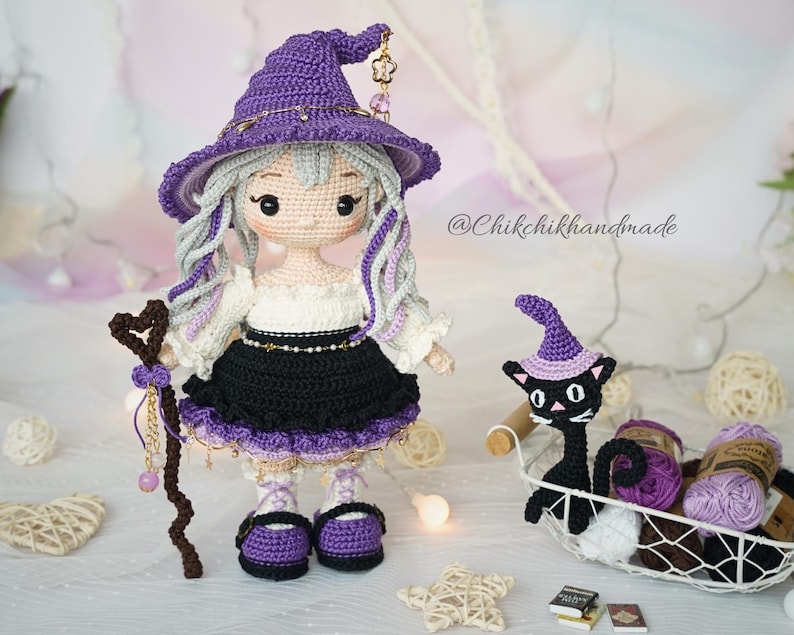 May include: A handcrafted crochet doll featuring light skin, long wavy silver hair, and a purple witch hat with gold charms. The doll wears a white blouse, black skirt, and purple shoes, holding a brown heart-topped staff. A small black cat wears a purple hat.