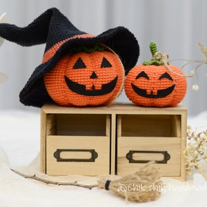 May include: Two crocheted pumpkins, one wearing a black witch's hat, sitting on a wooden shelf with two drawers. The pumpkins have orange bodies and black facial features. The smaller pumpkin is smiling, while the larger pumpkin has a more mischievous expression.