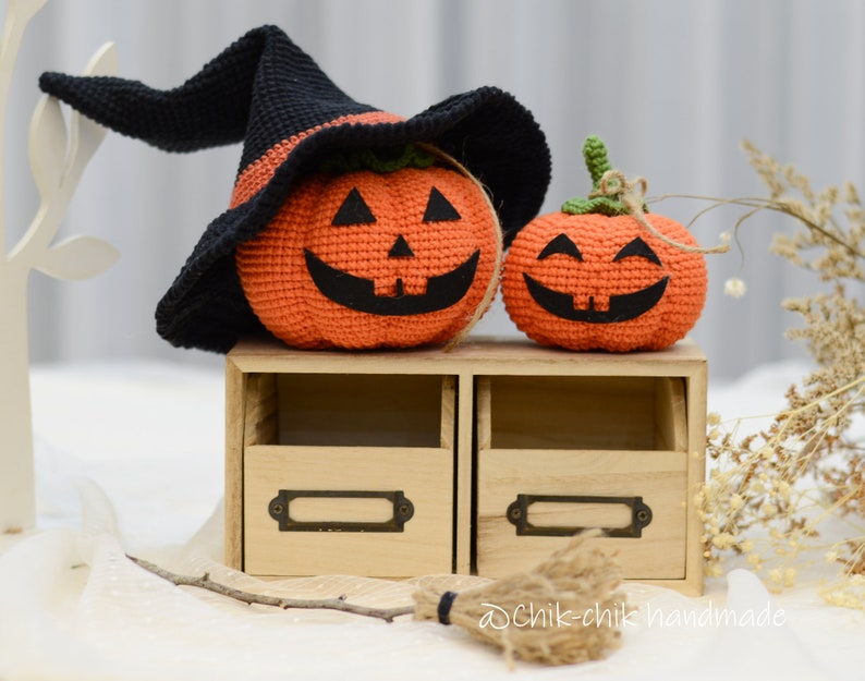 May include: Two crocheted pumpkins, one wearing a black witch's hat, sitting on a wooden shelf with two drawers. The pumpkins have orange bodies and black facial features. The smaller pumpkin is smiling, while the larger pumpkin has a more mischievous expression.