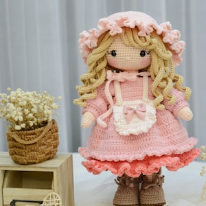 Pode incluir: Uma boneca de croch&ecirc; com cabelo loiro, vestindo um vestido rosa com um avental branco e sapatos marrons. A boneca est&aacute; em p&eacute; sobre uma superf&iacute;cie de madeira com uma pequena cesta de flores no fundo.