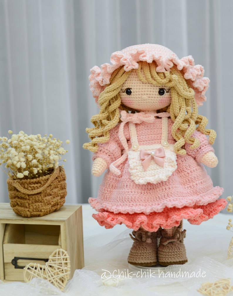 Pode incluir: Uma boneca de croch&ecirc; com cabelo loiro, vestindo um vestido rosa com um avental branco e sapatos marrons. A boneca est&aacute; em p&eacute; sobre uma superf&iacute;cie de madeira com uma pequena cesta de flores no fundo.