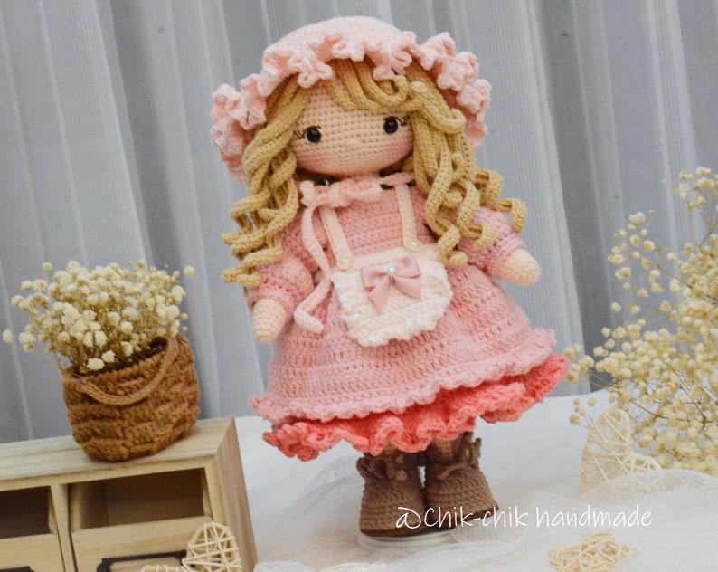 Pode incluir: Uma boneca de croch&ecirc; vestindo um vestido rosa com um avental branco e um la&ccedil;o rosa. A boneca tem cabelo longo e loiro e olhos castanhos. A boneca est&aacute; usando botas marrons. A boneca est&aacute; em p&eacute; em uma superf&iacute;cie branca. O texto "dChik-chik handmade" &eacute; vis&iacute;vel no canto inferior direito da imagem.