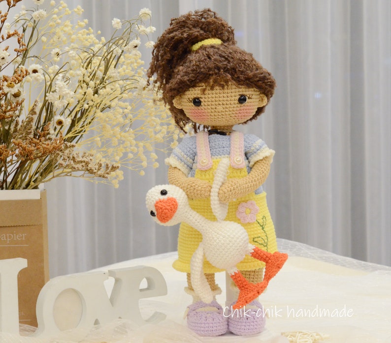 May include: A crocheted doll with brown hair in a ponytail, wearing a yellow dress with blue and pink accents, holding a white crocheted goose. The doll is standing on a white surface with the word "LOVE" in white letters.