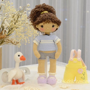 May include: A crocheted doll with brown hair, a blue and white striped shirt, denim shorts, and purple shoes. The doll is standing next to a crocheted goose and a yellow crocheted outfit with pink straps.