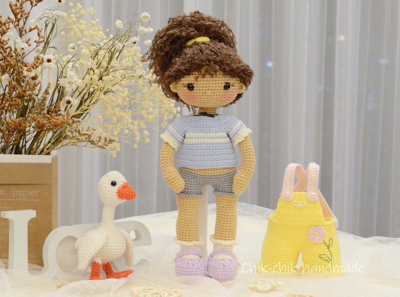 May include: A crocheted doll with brown hair, a blue and white striped shirt, denim shorts, and purple shoes. The doll is standing next to a crocheted goose and a yellow crocheted outfit with pink straps.