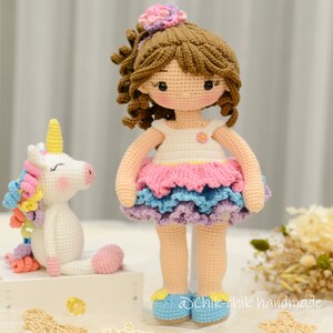 May include: A crocheted doll with brown hair and a pink and blue dress stands next to a crocheted unicorn with a rainbow mane. The doll is wearing blue shoes and has a flower in her hair. The unicorn is white with a pink nose and closed eyes. The text "@chik-chik handmade" is visible in the bottom right corner of the image.