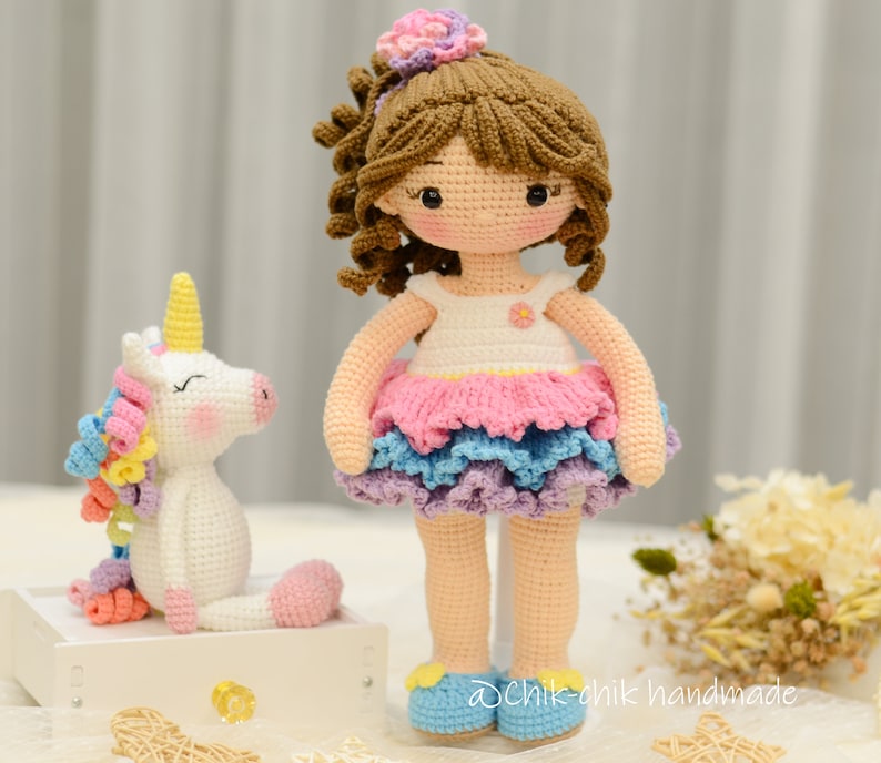 May include: A crocheted doll with brown hair and a pink and blue dress stands next to a crocheted unicorn with a rainbow mane. The doll is wearing blue shoes and has a flower in her hair. The unicorn is white with a pink nose and closed eyes. The text "@chik-chik handmade" is visible in the bottom right corner of the image.