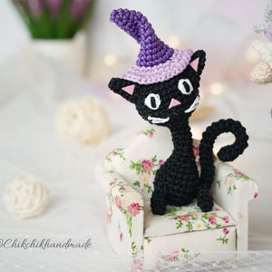 May include: A black crocheted cat figurine wearing a purple witch hat with a light purple brim. The cat has white eyes, a pink nose, and white whiskers. It is sitting on a small armchair with a white base and a floral pattern of pink and green.
