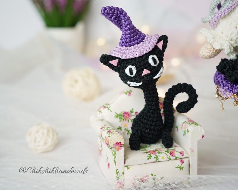May include: A black crocheted cat figurine wearing a purple witch hat with a light purple brim. The cat has white eyes, a pink nose, and white whiskers. It is sitting on a small armchair with a white base and a floral pattern of pink and green.