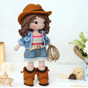 K&ouml;nnte beinhalten: Geh&auml;kelte Puppe mit Jeansjacke, buntem Tanktop, Jeans-Shorts und braunen Cowboy-Stiefeln. Die Puppe h&auml;lt ein braunes Seil und tr&auml;gt einen braunen Cowboyhut.