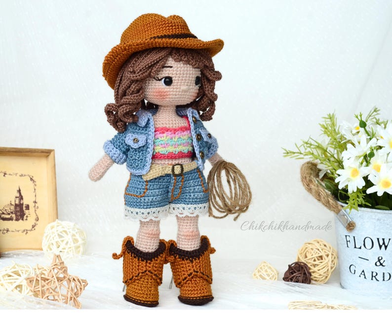 K&ouml;nnte beinhalten: Geh&auml;kelte Puppe mit Jeansjacke, buntem Tanktop, Jeans-Shorts und braunen Cowboy-Stiefeln. Die Puppe h&auml;lt ein braunes Seil und tr&auml;gt einen braunen Cowboyhut.