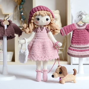 May include: A crocheted doll with long blonde hair wearing a pink dress and a burgundy hat. The doll is holding a white crocheted bag and a lead attached to a brown crocheted dachshund. The doll is standing on a white base. There are two other crocheted garments on hangers, a brown jacket and a pink dress.