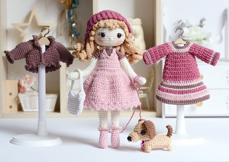 May include: A crocheted doll with long blonde hair wearing a pink dress and a burgundy hat. The doll is holding a white crocheted bag and a lead attached to a brown crocheted dachshund. The doll is standing on a white base. There are two other crocheted garments on hangers, a brown jacket and a pink dress.
