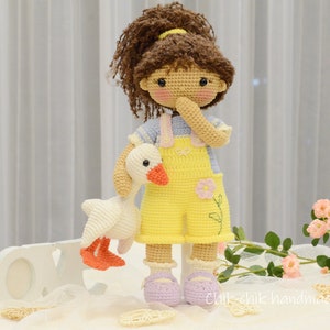 May include: A crocheted doll with brown hair wearing a yellow overall dress and a blue and white striped shirt. The doll is holding a white crocheted goose. The doll is standing on a white surface with a white wooden sign that says "LOVE" in front of it.