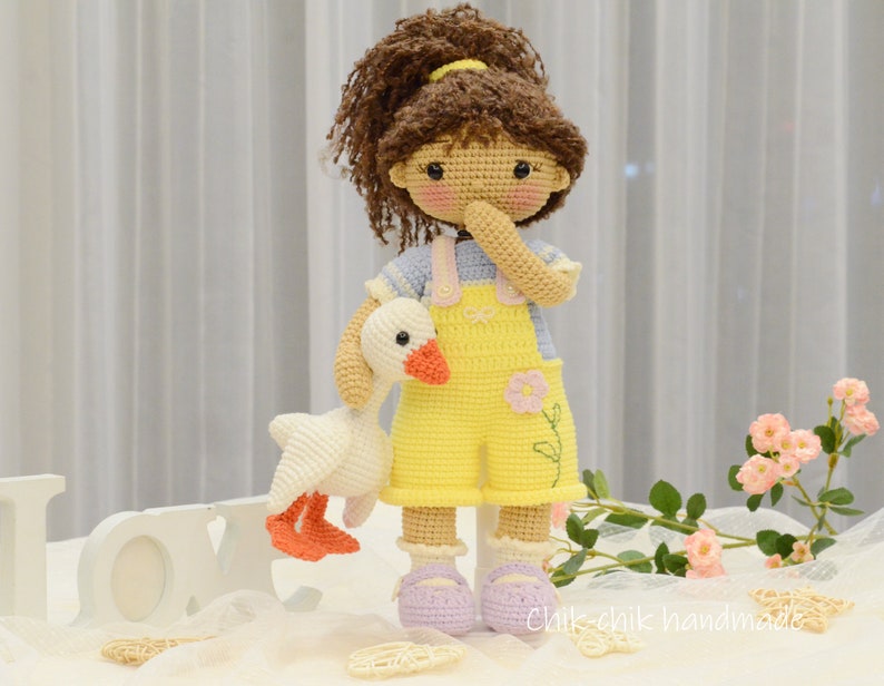 May include: A crocheted doll with brown hair wearing a yellow overall dress and a blue and white striped shirt. The doll is holding a white crocheted goose. The doll is standing on a white surface with a white wooden sign that says "LOVE" in front of it.