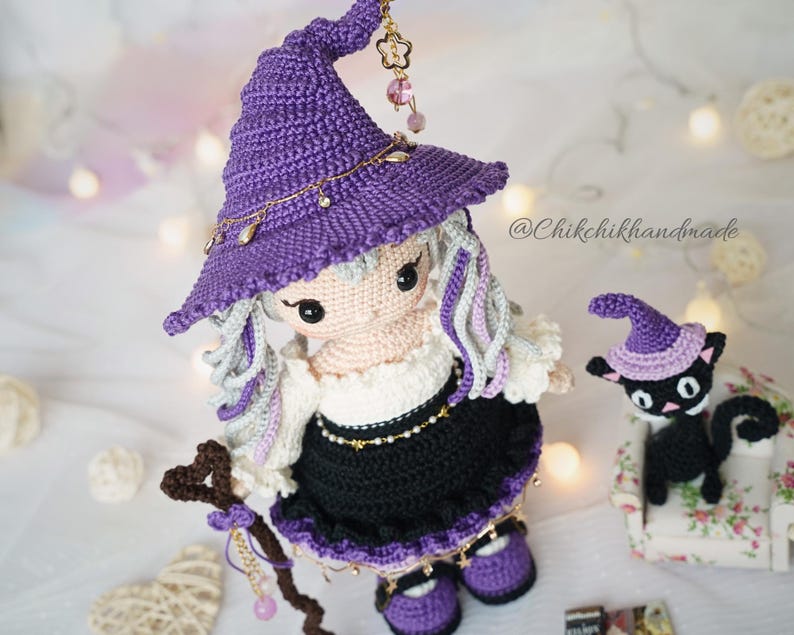 May include: A handcrafted crochet doll wearing a purple pointed hat with gold charms, a white blouse, and a black skirt, holding a brown staff. Beside the doll is a small black crochet cat wearing a matching purple hat. The text @Chikchikhandmade is visible.