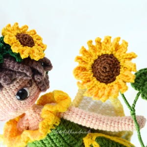 May include: A crocheted doll with brown hair wearing a sunflower crown and a green and yellow dress. The doll is holding a crocheted sunflower with green leaves.
