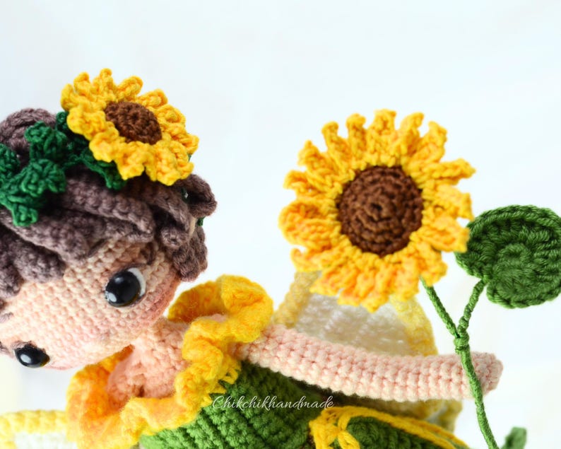 May include: A crocheted doll with brown hair wearing a sunflower crown and a green and yellow dress. The doll is holding a crocheted sunflower with green leaves.