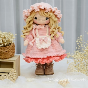 Pode incluir: Uma boneca de croch&ecirc; com cabelo loiro, vestindo um vestido rosa com um avental branco e um la&ccedil;o rosa. A boneca est&aacute; usando sapatos marrons e tem um chap&eacute;u rosa de croch&ecirc;.