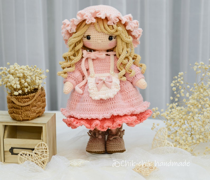 Pode incluir: Uma boneca de croch&ecirc; com cabelo loiro, vestindo um vestido rosa com um avental branco e um la&ccedil;o rosa. A boneca est&aacute; usando sapatos marrons e tem um chap&eacute;u rosa de croch&ecirc;.