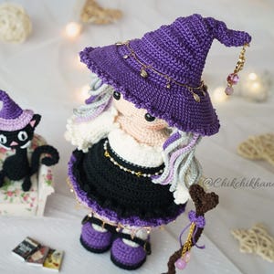 May include: A crochet doll wearing a purple pointed hat with gold-toned charms, a white top, and a black skirt, featuring long wavy hair. Beside the doll is a small black crochet cat wearing a purple hat, sitting on a miniature floral armchair.