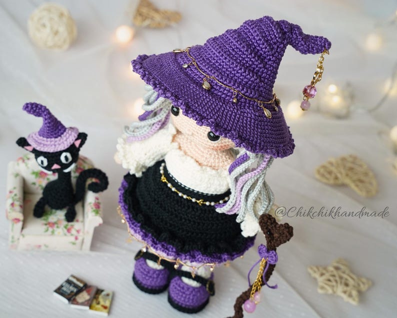 May include: A crochet doll wearing a purple pointed hat with gold-toned charms, a white top, and a black skirt, featuring long wavy hair. Beside the doll is a small black crochet cat wearing a purple hat, sitting on a miniature floral armchair.