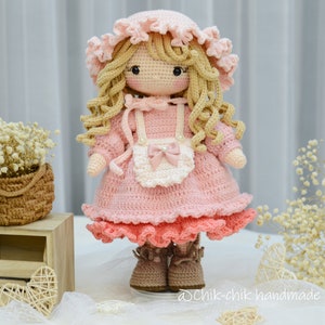 Pode incluir: Uma boneca de croch&ecirc; vestindo um vestido rosa com um avental branco e um la&ccedil;o rosa. A boneca tem cabelo loiro encaracolado e est&aacute; usando sapatos marrons. A boneca est&aacute; em p&eacute; em uma superf&iacute;cie branca com uma cesta de flores e uma caixa de madeira no fundo.