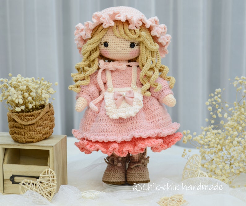 Pode incluir: Uma boneca de croch&ecirc; vestindo um vestido rosa com um avental branco e um la&ccedil;o rosa. A boneca tem cabelo loiro encaracolado e est&aacute; usando sapatos marrons. A boneca est&aacute; em p&eacute; em uma superf&iacute;cie branca com uma cesta de flores e uma caixa de madeira no fundo.