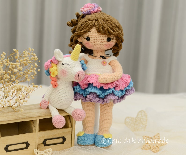May include: A crocheted doll with brown hair and a pink, blue, and purple dress holds a white crocheted unicorn with a rainbow mane. The doll is wearing a white shirt with a flower design. The doll and unicorn are sitting on a wooden shelf with drawers.