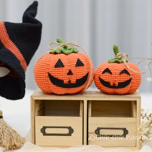 May include: Two crocheted orange pumpkins with black eyes and mouths, one larger than the other, sitting on a wooden shelf with two drawers. The smaller pumpkin has a happy expression, while the larger pumpkin has a more mischievous expression. The pumpkins are decorated with green stems and brown twine.