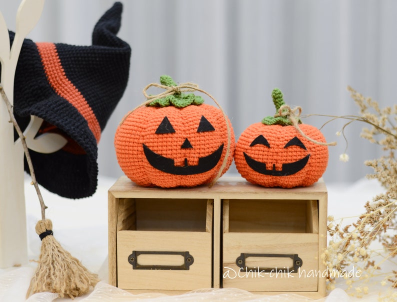 May include: Two crocheted orange pumpkins with black eyes and mouths, one larger than the other, sitting on a wooden shelf with two drawers. The smaller pumpkin has a happy expression, while the larger pumpkin has a more mischievous expression. The pumpkins are decorated with green stems and brown twine.