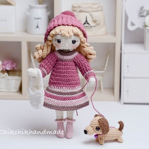 May include: A crocheted doll wearing a pink dress, hat, and jumper, holding a white crocheted bag and a lead attached to a brown crocheted dachshund. The doll has blonde hair and brown eyes. The image includes the text "@Chikchikhandmade".