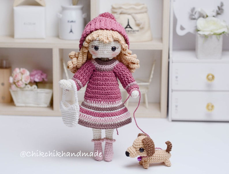 May include: A crocheted doll wearing a pink dress, hat, and jumper, holding a white crocheted bag and a lead attached to a brown crocheted dachshund. The doll has blonde hair and brown eyes. The image includes the text "@Chikchikhandmade".