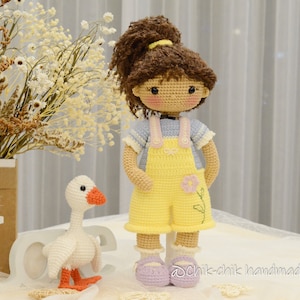 May include: A crocheted doll with brown hair, wearing a blue and white striped shirt and yellow overalls with a flower design. The doll is standing next to a white crocheted goose with orange feet.