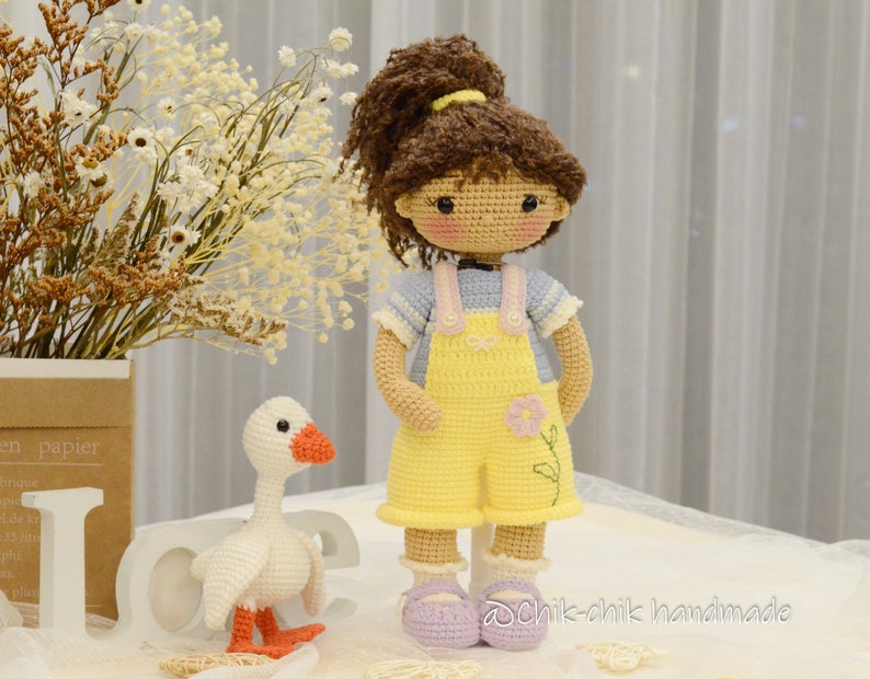 May include: A crocheted doll with brown hair, wearing a blue and white striped shirt and yellow overalls with a flower design. The doll is standing next to a white crocheted goose with orange feet.