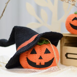 May include: Two crocheted pumpkins, one wearing a black witch hat with an orange brim. The pumpkins have black eyes, a nose, and a wide smile. The pumpkins are sitting on a wooden shelf with a label that says "@Chik-chik handmade".