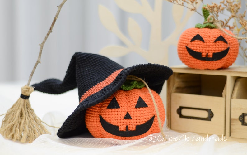 May include: Two crocheted pumpkins, one wearing a black witch hat with an orange brim. The pumpkins have black eyes, a nose, and a wide smile. The pumpkins are sitting on a wooden shelf with a label that says "@Chik-chik handmade".