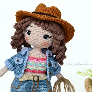 K&ouml;nnte beinhalten: Eine geh&auml;kelte Puppe mit einem braunen Cowboyhut, einer blauen Jeansjacke, einem bunten gestreiften Oberteil, blauen Jeans und einem braunen G&uuml;rtel. Die Puppe hat braunes lockiges Haar und h&auml;lt ein Seil.