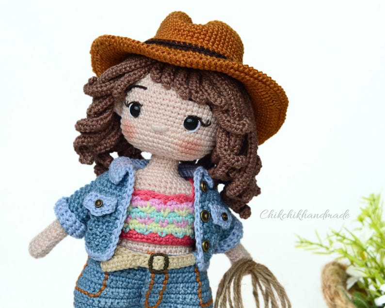 K&ouml;nnte beinhalten: Eine geh&auml;kelte Puppe mit einem braunen Cowboyhut, einer blauen Jeansjacke, einem bunten gestreiften Oberteil, blauen Jeans und einem braunen G&uuml;rtel. Die Puppe hat braunes lockiges Haar und h&auml;lt ein Seil.