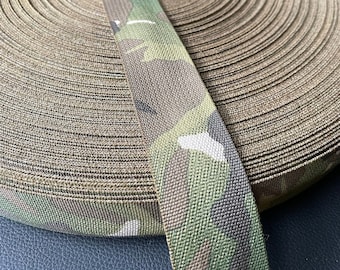 May include: A roll of camouflage webbing with a green, brown, and tan pattern. The webbing is rolled up tightly and is about 1 inch wide.