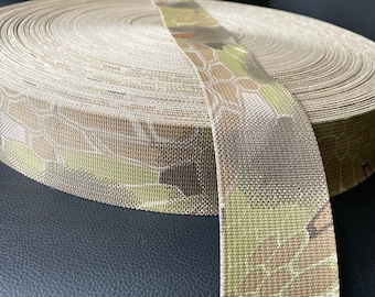 May include: A roll of camouflage webbing featuring a repeating pattern of green, brown, and tan geometric shapes. The webbing is tightly wound, with a section unrolled and laid across the top. The material appears to be durable and suitable for outdoor use.