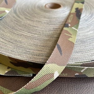Peut inclure: Un rouleau de sangle de camouflage avec un motif brun, vert et noir. La sangle est enroulée et le bout du rouleau est visible.