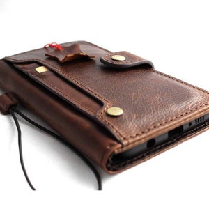 Könnte beinhalten: Eine dunkelbraune Handy-Geldbörsenhülle aus Leder mit strukturierter Oberfläche, Druckknopfverschluss und einem externen Kartenfach. Die Hülle enthält ein kleines Lederetikett, das mit einer roten Schnur befestigt ist, sowie eine dünne schwarze Handschlaufe.