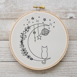 Puede incluir: Un aro de bordado blanco con un dibujo de línea negra de un gato sentado en una luna creciente con estrellas y una bola de hilo colgante. El gato está rodeado de una corona de flores.